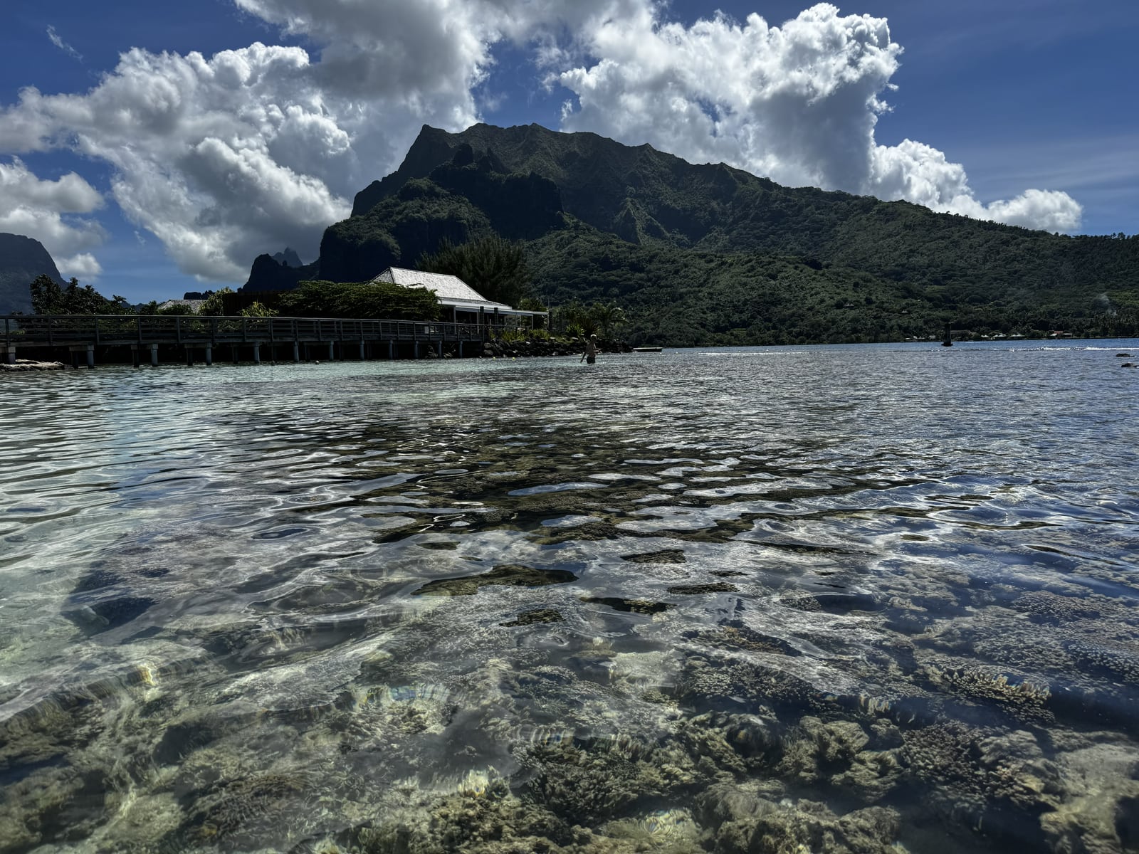 Clear shallow water with visible stones in the foreground and a green mountain ridge beneath large clouds.