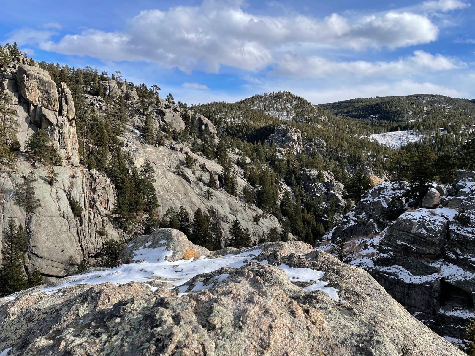 Granite outcrops and patches of snow among pines, with forested hills under a bright sky.