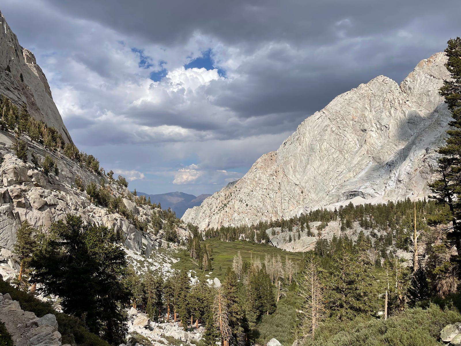 A forested valley between a dark cliff and a pale granite ridge, under heavy grey clouds with a patch of blue sky.