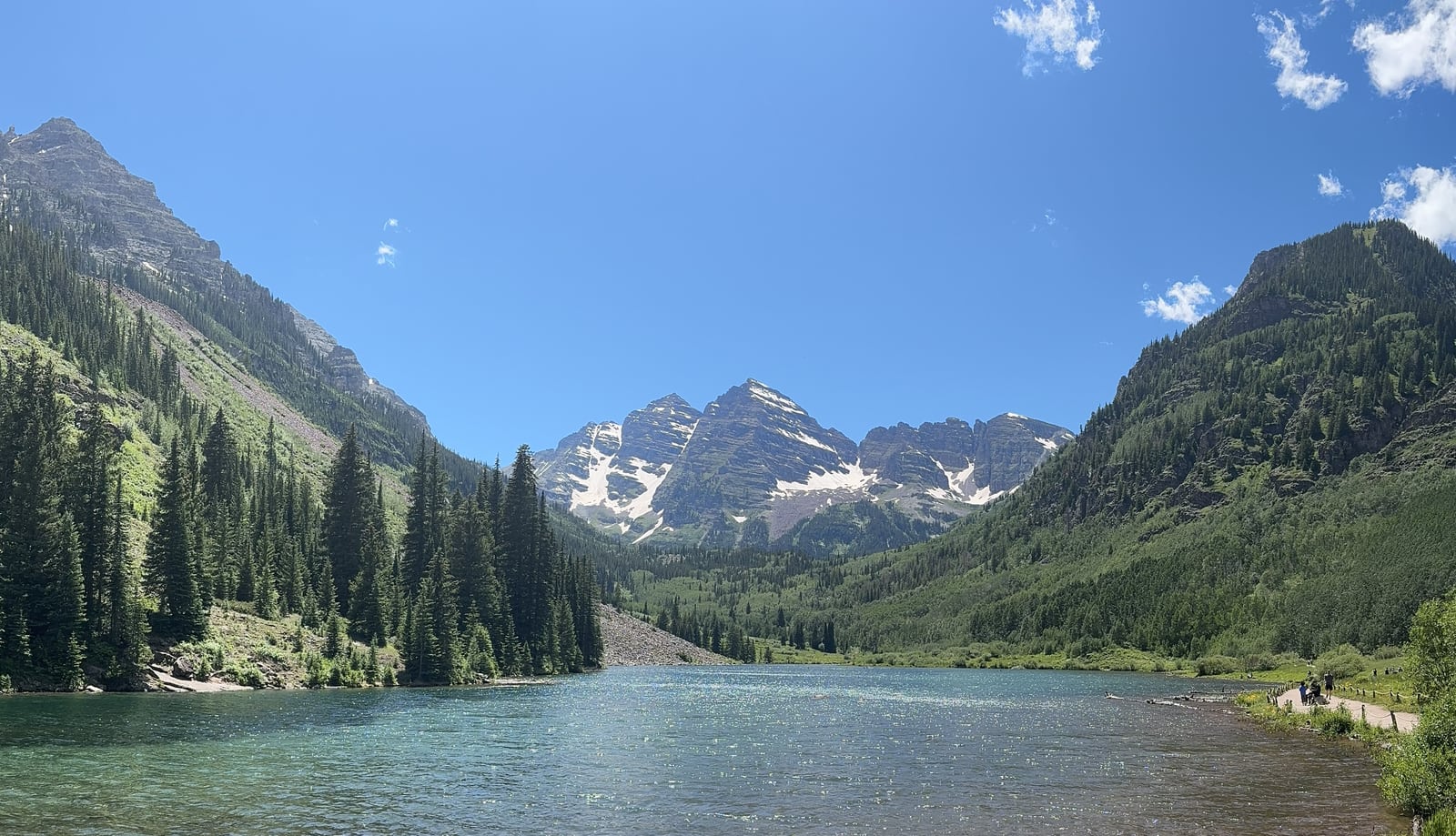 A mountain lake between pine-covered slopes, with snow-streaked peaks beneath a clear blue sky.