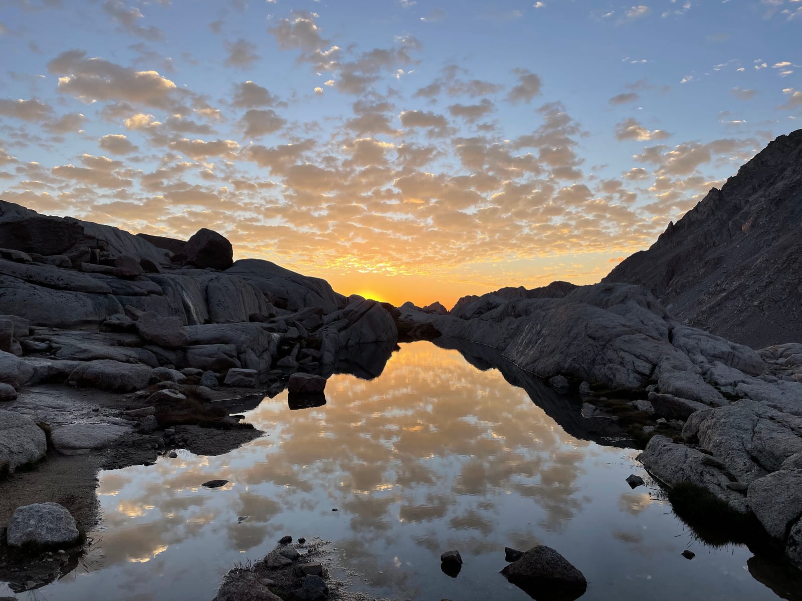 Golden light and clouds reflected in a still pool between rocky mountain slopes.