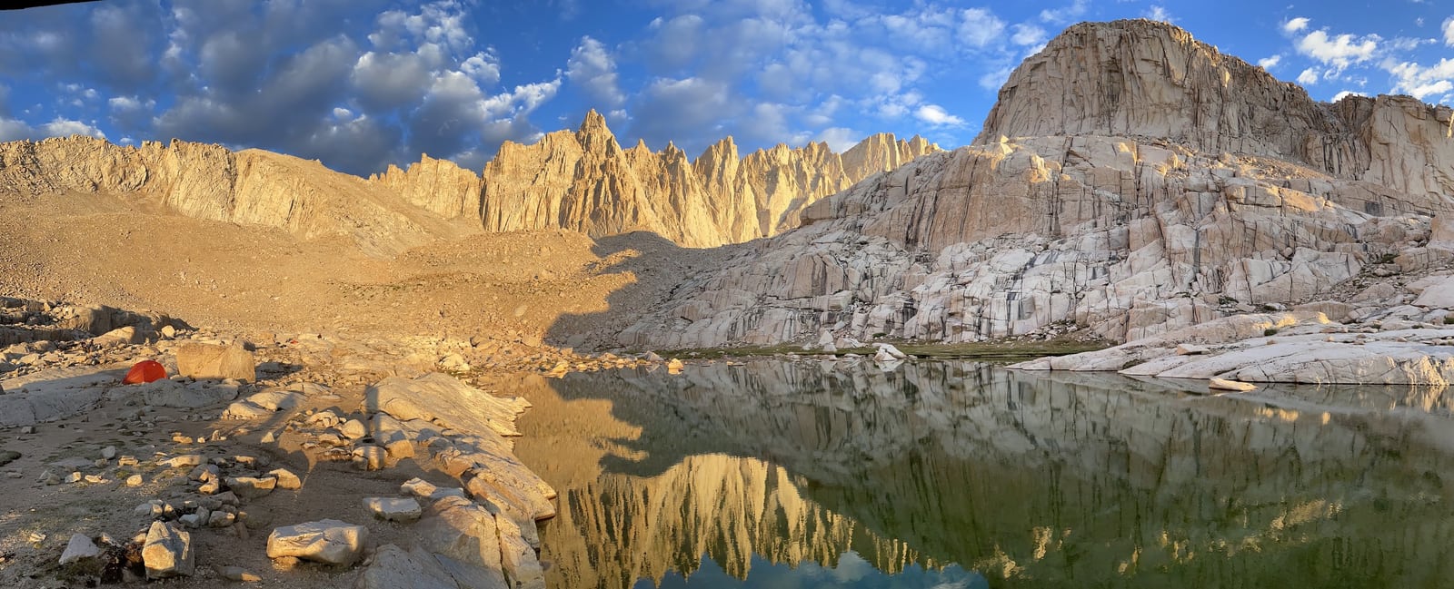 Sunlit rocky ridgelines reflected in a still lake, with a small red tent on the shore.