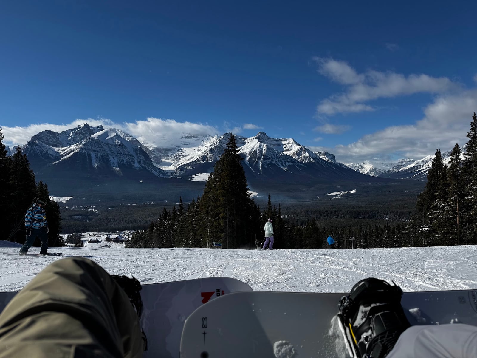 A snowy ski slope below distant snow-covered mountains, with a snowboard and resting legs in the foreground.